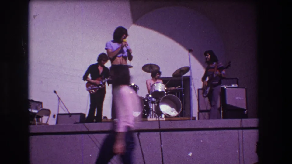 1973 Medford, New Jersey 8mm frame with a blurred passerby in the foreground and the band performing on the raised stage.
