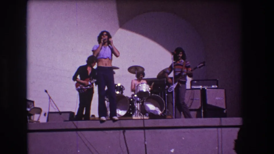 1973 Medford, New Jersey frame showing the band in a wider stage arrangement with singer, drummer, guitarist, bassist, amps, and backdrop.