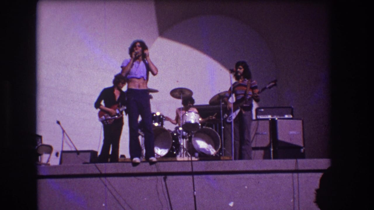 Rock band performs on stage with singer, guitarists, drummer, amplifiers, and curved backdrop.