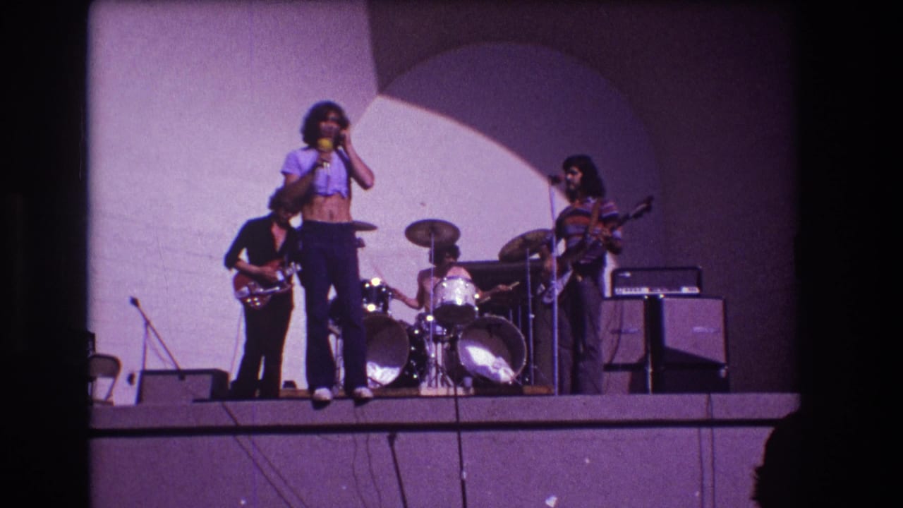 Band continues performing on stage with singer at center, drummer behind, and guitarists on both sides.