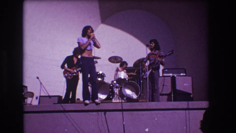 1973 Medford, New Jersey frame showing the vocalist in profile with guitarists, drummer, drum kit, cables, and amplifiers visible.