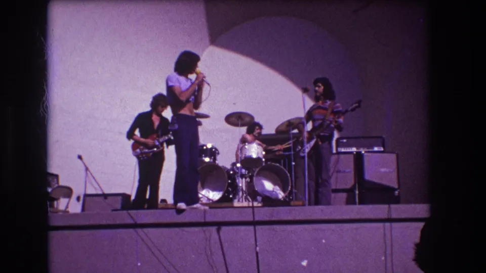1973 Medford stage frame showing the singer turned toward a guitarist, with drummer, amplifiers, and curved backdrop in view.