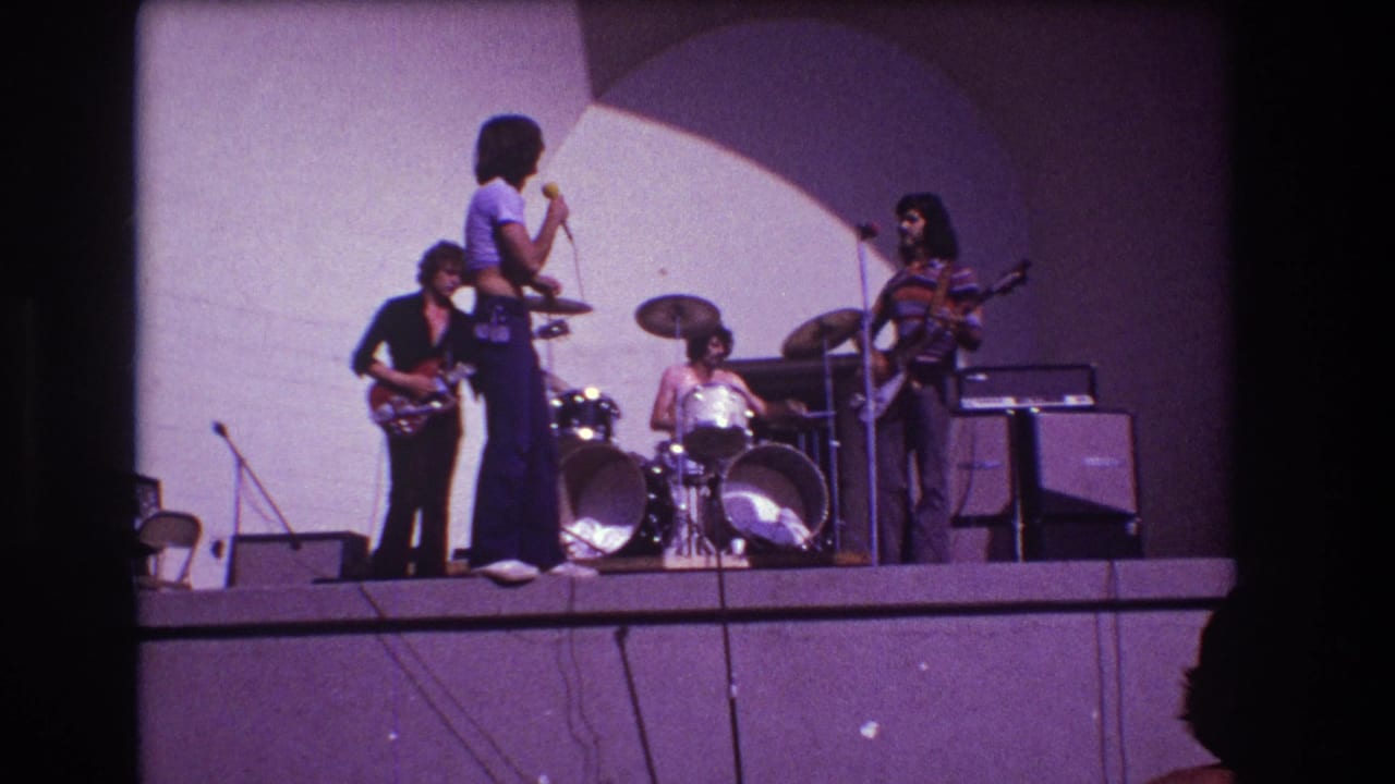 1973 Medford, New Jersey 8mm frame showing a rock band performing on a raised stage with a standing singer, two guitarists, drummer, microphone stands, and amplifiers.