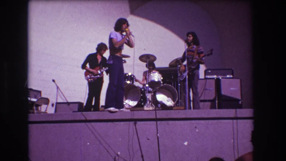 1973 Medford, New Jersey 8mm frame showing the lead singer near center stage, backed by a guitarist, drummer, second guitarist, drum kit, and amplifiers.