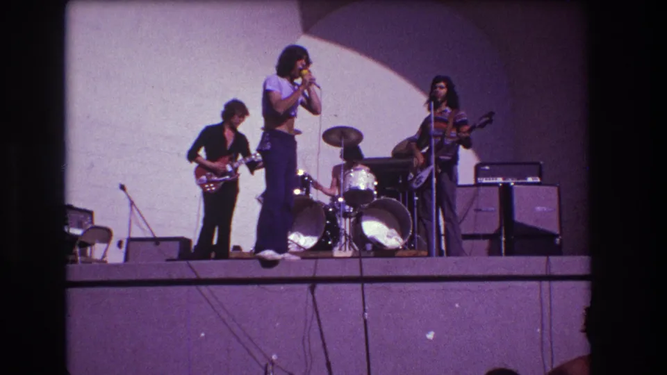 1973 Medford, New Jersey 8mm frame of a local rock band continuing its set, with singer at microphone, two guitarists, drummer, drum kit, amplifiers, and stage cables visible.