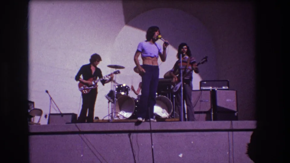 1973 Medford, New Jersey 8mm still of a singer in a cropped purple shirt at center stage with band members and equipment around him.