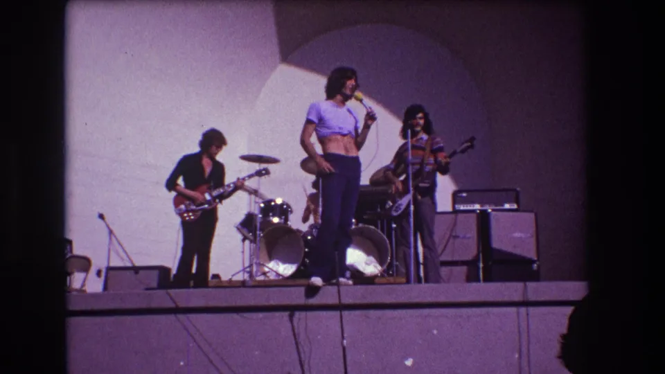 1973 Medford, New Jersey frame showing the singer and bassist near microphones, with guitar, drums, amplifiers, and a simple stage backdrop.