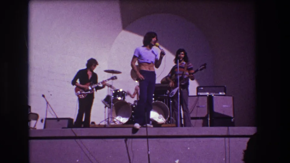 1973 Medford, New Jersey 8mm frame showing a rock band on a raised stage with a lead singer at a microphone, guitarist, bassist, drum kit, and amplifiers.
