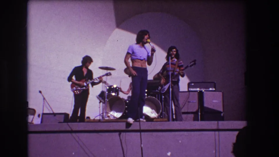 1973 Medford, New Jersey frame of the band mid-performance, with the singer holding the microphone while guitar, bass, drums, and stage amplifiers remain visible.