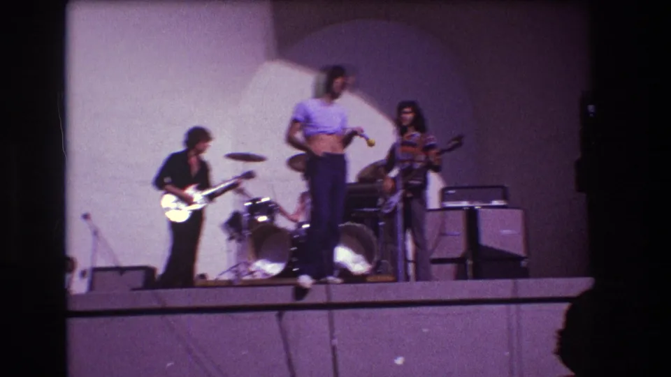 Soft 8mm frame from the 1973 Medford performance showing the singer standing near center as the guitarist, drummer, bassist, and amplifiers remain in view.