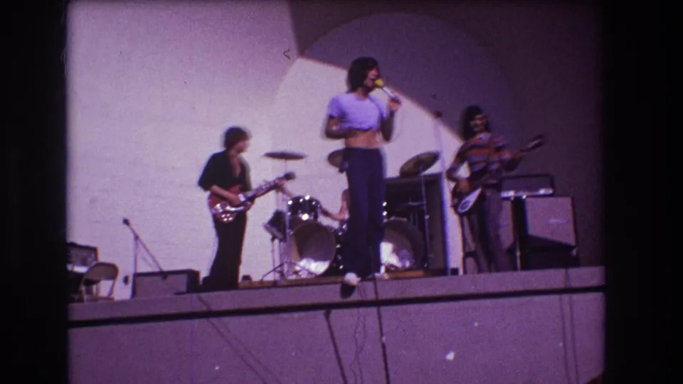 1973 Medford, New Jersey stage frame with the lead singer turned toward the band, guitarist at left, drummer behind, bassist at right, and amplifiers along the backline.