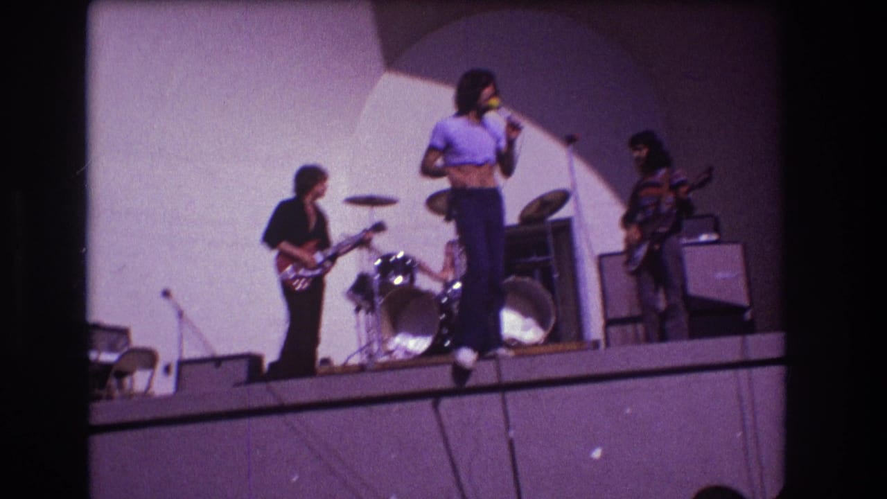 Rock band performs on a raised stage with singer, guitarists, drum kit, amplifiers, and pale backdrop.