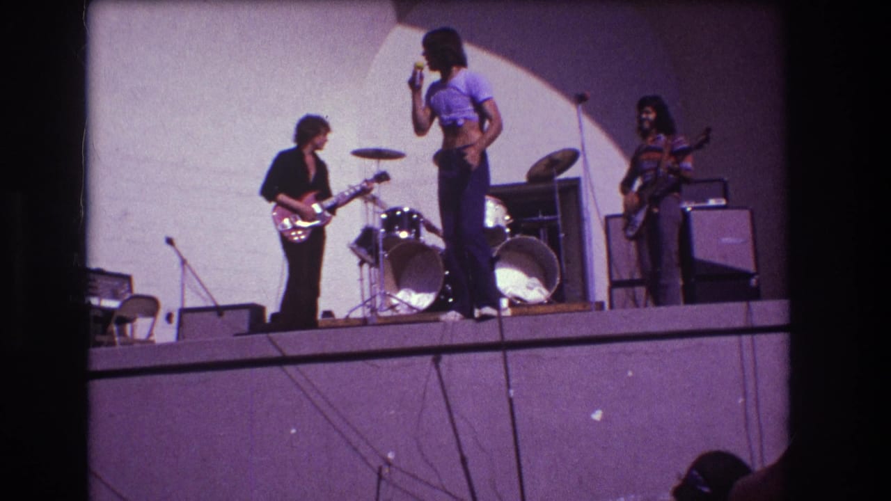 Singer faces a guitarist while the drummer, second guitarist, amplifiers, and stage cables are visible.