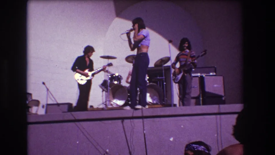 1973 Medford 8mm home-movie frame of a live band on a raised stage, with singer, guitarists, drums, and amps under purple-tinted color.
