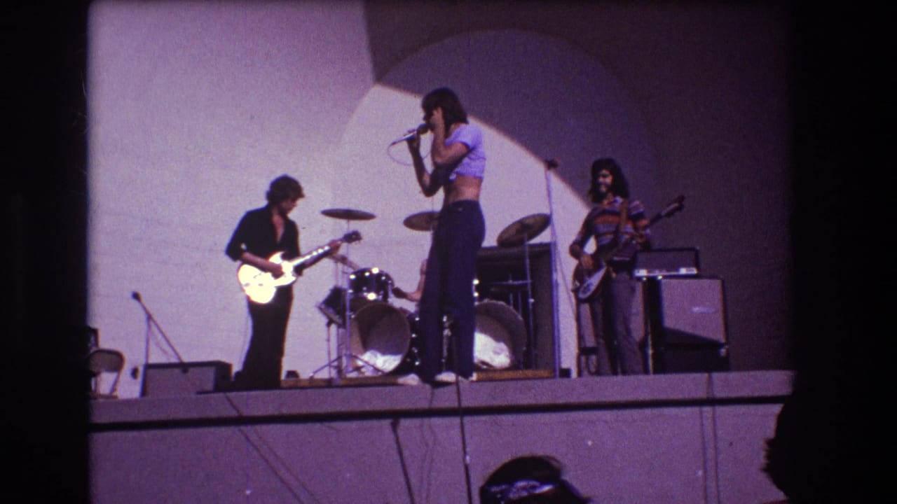 Rock band on stage with vocalist at microphone, guitarist, bassist, drum kit, and amplifiers.