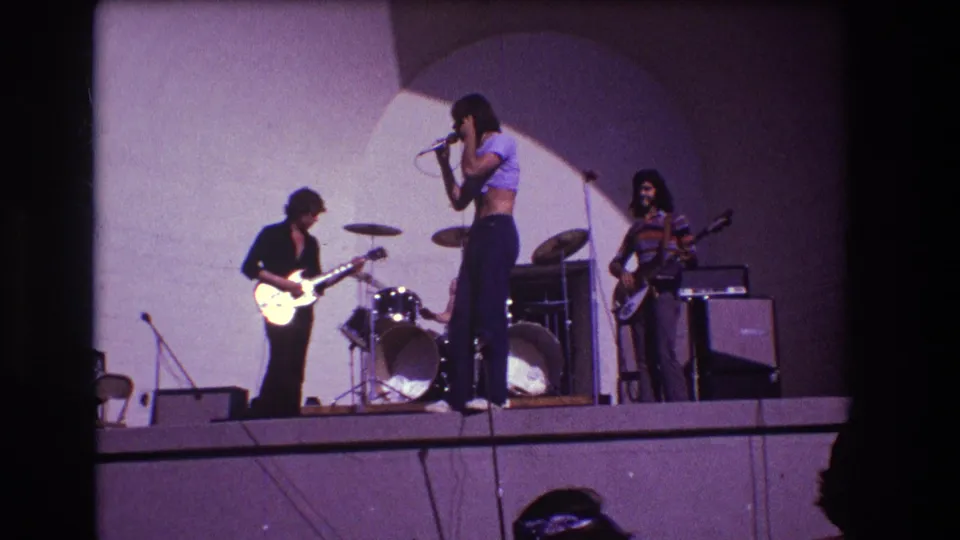 1973 Medford, New Jersey home-movie frame showing a rock band warming up on stage with singer, guitarist, bassist, drums, and amps.