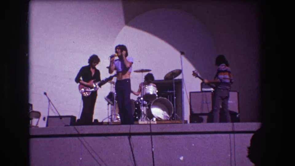 1973 Medford, New Jersey 8mm frame of a singer in a purple cropped top performing with guitarist, drummer, and bassist on stage.