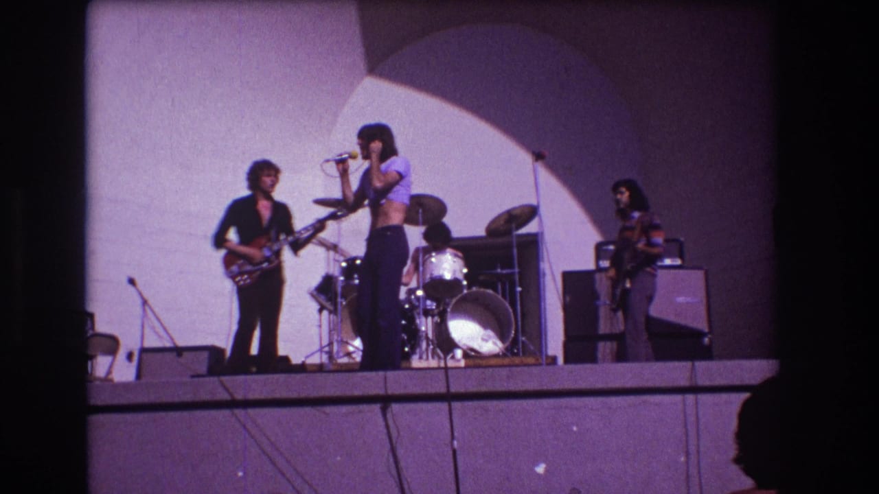 1973 Medford, New Jersey 8mm frame showing a singer at a microphone, guitarist, drummer, and bassist on a simple stage.