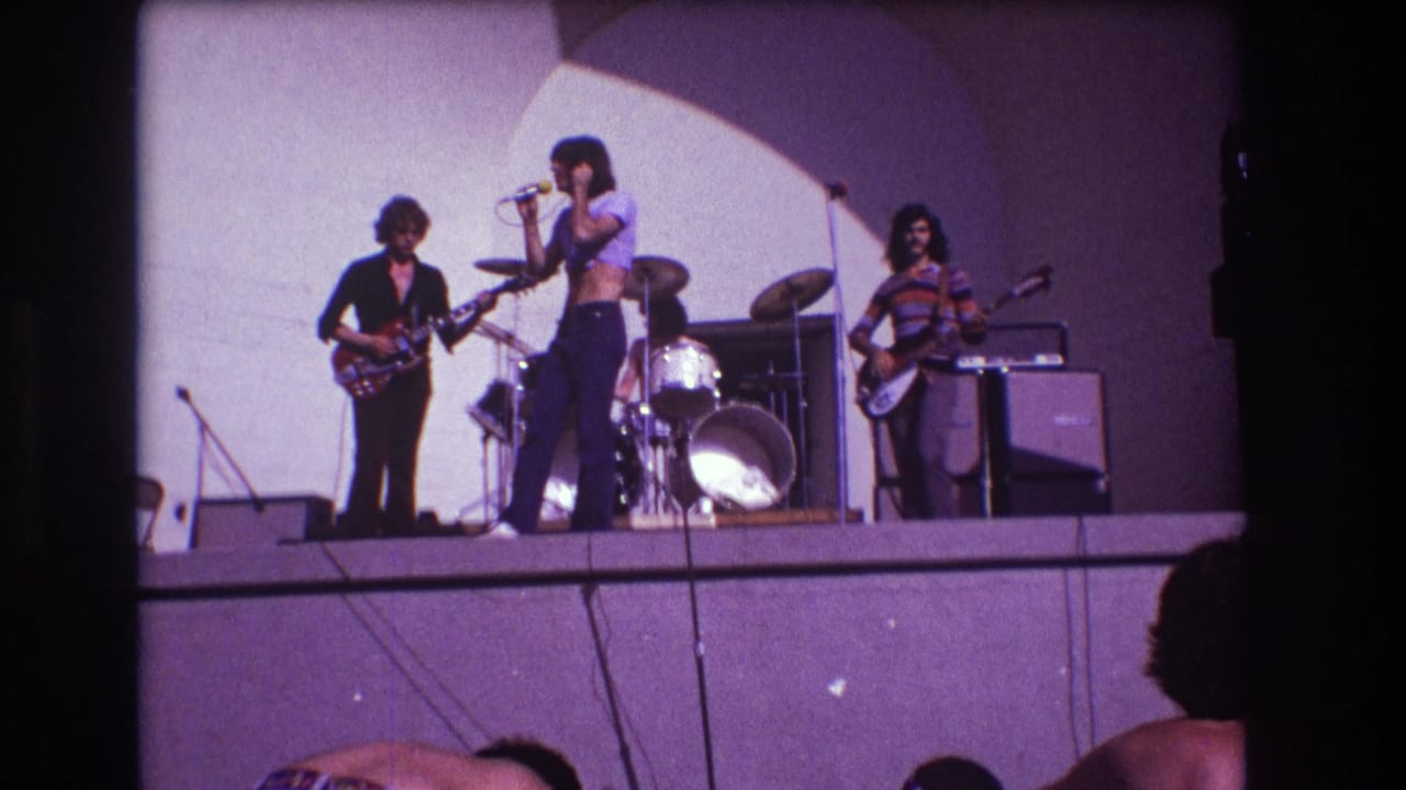 Audience-shot 1973 Medford concert frame with a central singer, two guitarists, drum kit, amplifiers, and white arched backdrop.