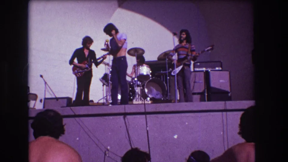1973 Medford 8mm still of a rock band mid-performance, with singer, guitarist, drummer, bassist, amplifiers, and audience heads visible.