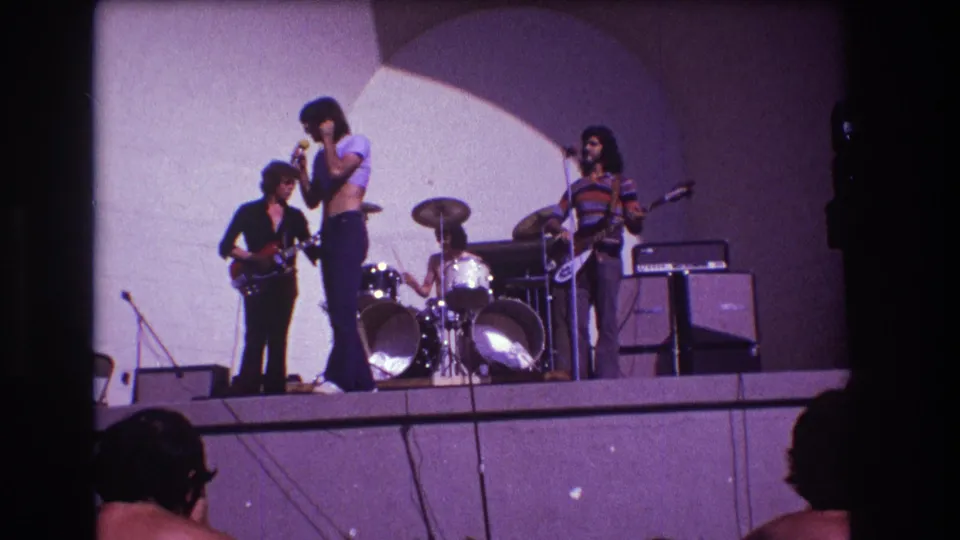 1973 Medford, New Jersey home-movie frame of a four-piece rock band performing before visible audience silhouettes.