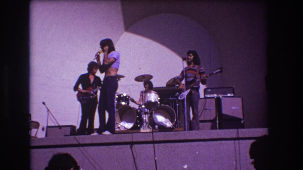 Singer stands at a microphone while band members play guitar, drums, and bass on stage.