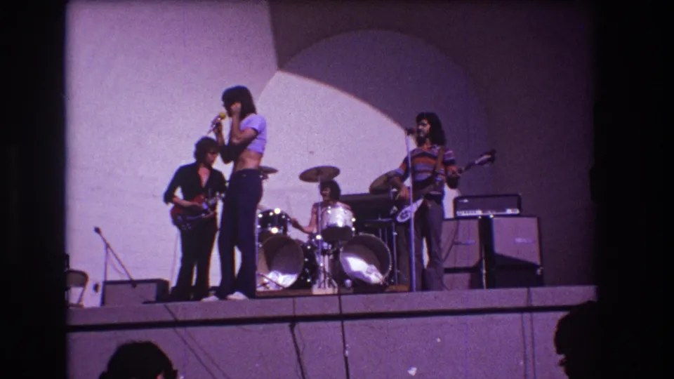 Audience-shot 1973 Medford concert frame with singer at center, drummer behind, and guitarists on either side under purple stage light.