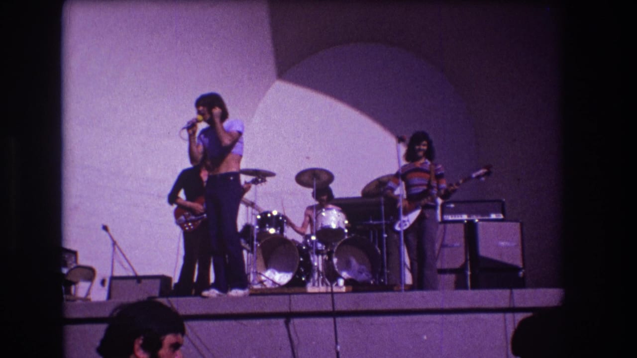 Rock band on stage with singer at microphone, guitarists, drummer, amplifiers, and audience heads below.