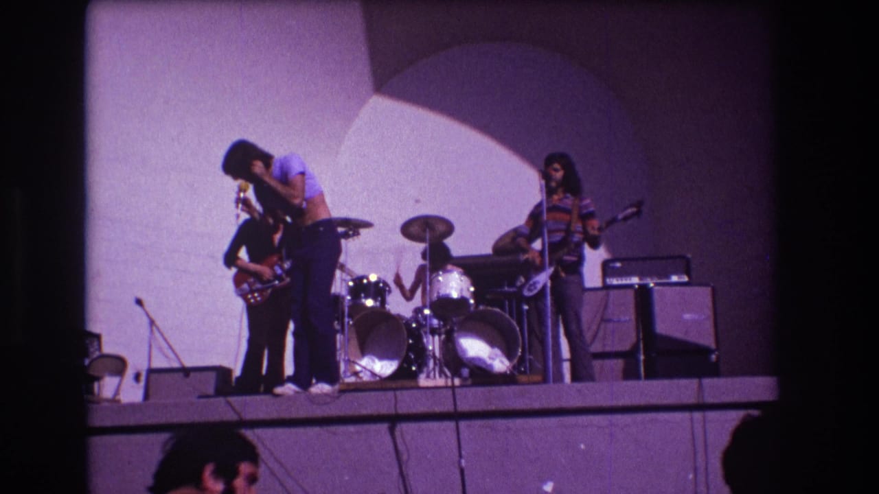 Singer leans toward a guitarist while drummer and striped-shirt guitarist continue playing on stage.