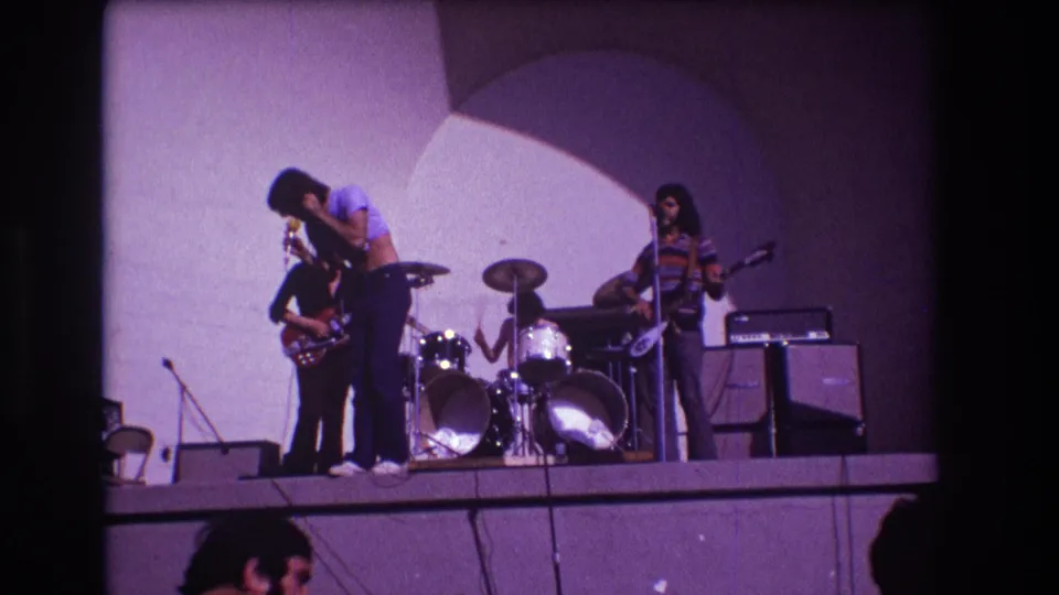 1973 Medford, New Jersey frame showing the singer bent toward a guitarist during a rock band performance on a lit stage.