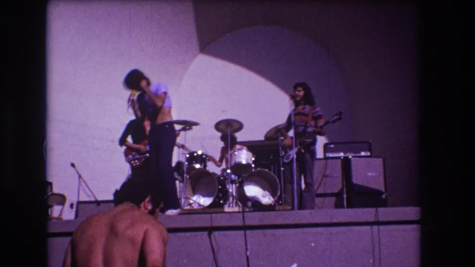 1973 Medford, New Jersey home-movie frame with singer, guitarists, drummer, amplifiers, and a nearby audience member in front.