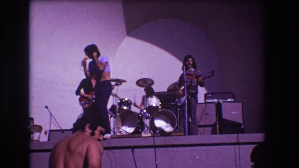 1973 Medford, New Jersey frame of a rock band mid-performance with purple stage color and audience figures along the bottom edge.