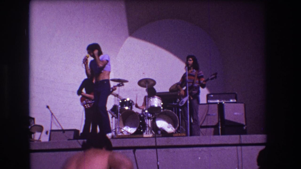Band on stage with a singer at a microphone, guitarists, drummer and audience silhouettes.