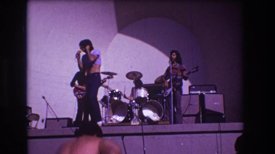 1973 Medford, New Jersey 8mm frame showing a Black male singer at a microphone with guitarists and drummer on a small stage.