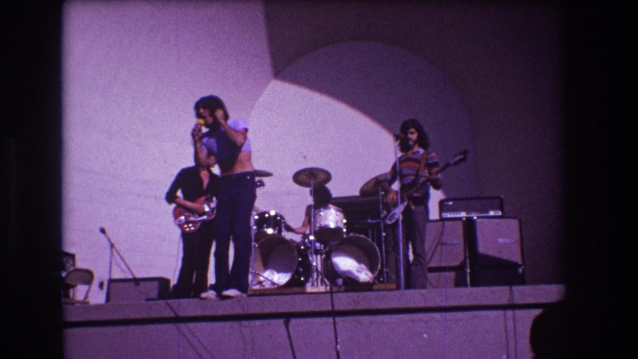 Singer faces left at a microphone as guitarists and drummer perform behind him.