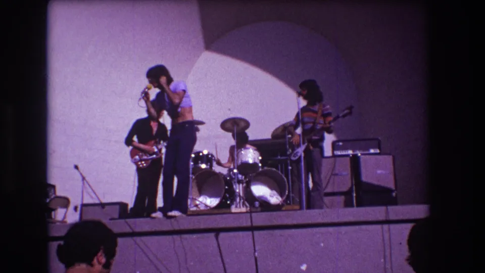 Audience-shot 8mm frame from Medford in 1973 showing a singer at center stage with guitar and drum accompaniment.
