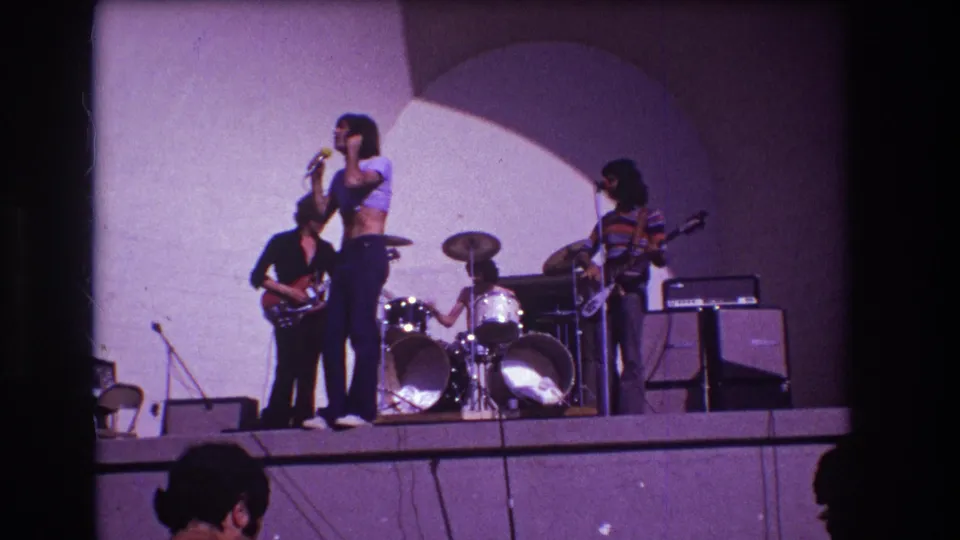 1973 Medford, New Jersey home-movie still of a singer performing with guitarists, drummer and stage equipment visible.