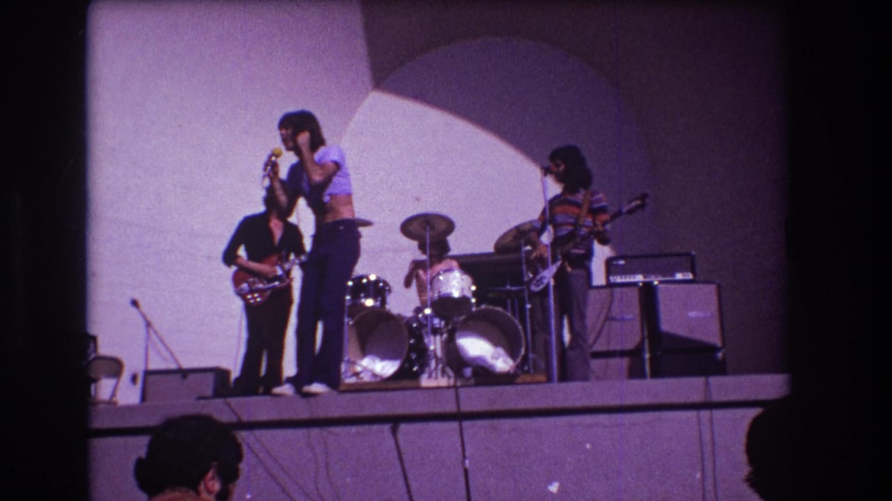 Four-piece band performs on a raised stage with singer at center, guitarists at sides, and drummer behind.