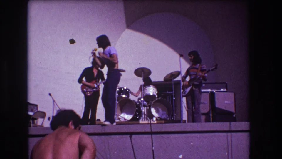 1973 Medford, New Jersey home-movie frame showing the singer turned toward a guitarist as the drummer and bassist perform nearby.