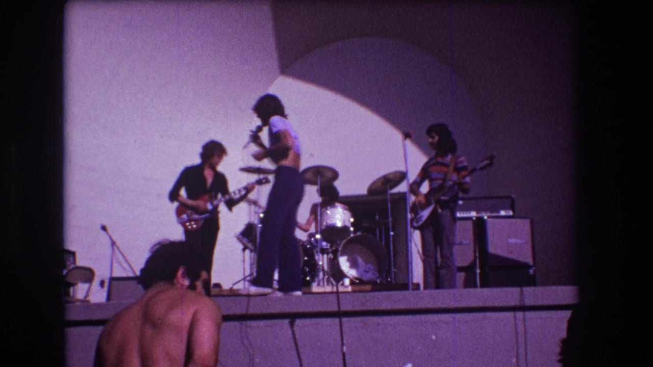 Rock band on a stage with singer, guitarist, drummer, bassist, amps, and audience foreground.