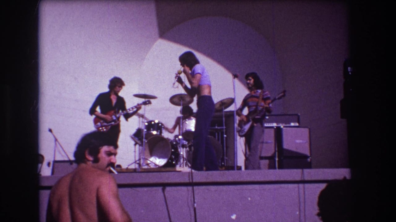 Singer stands near the microphone while guitar, bass, drums, amps, and audience figures are visible.