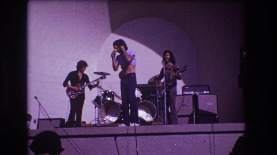 1973 Medford, New Jersey frame of a rock singer facing the audience as bandmates play guitar, drums, and bass on a small stage.