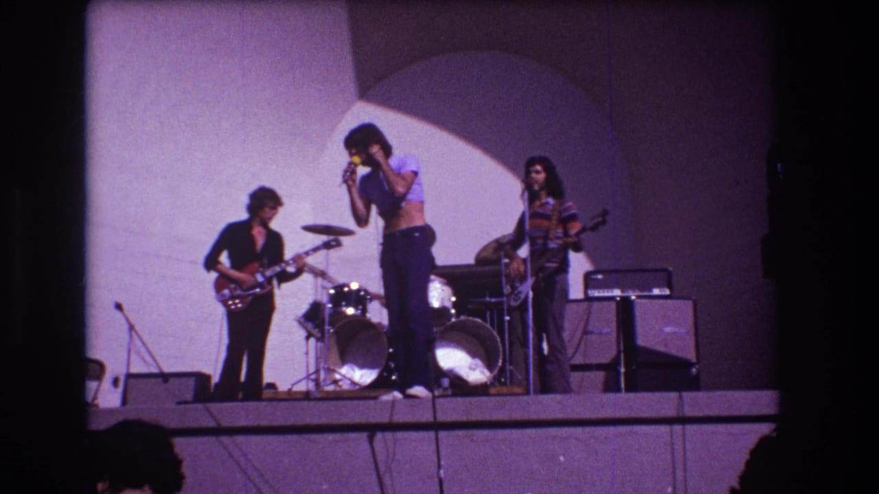 Three-piece rock band performs on a stage with drums, amplifiers, microphone stands, and an arched backdrop.