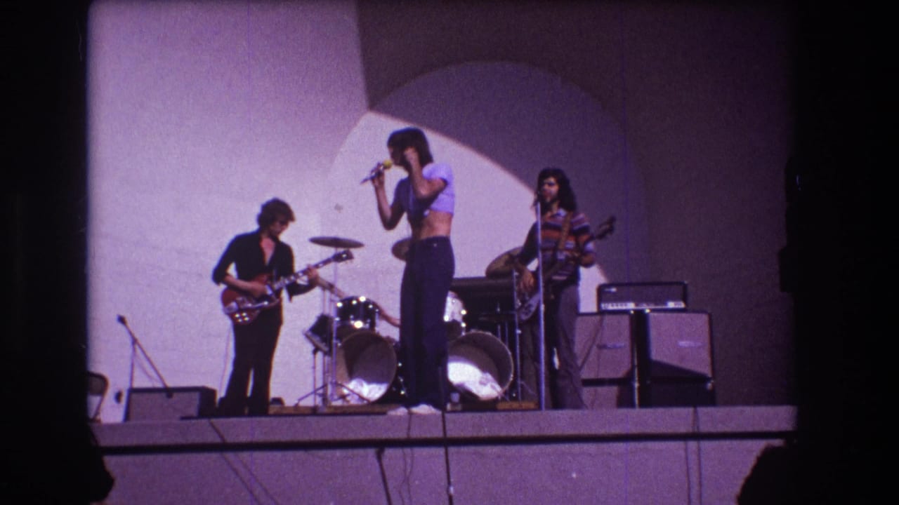 Lead singer holds a microphone while guitarist and bassist play beside a drum kit and amplifier cabinets.