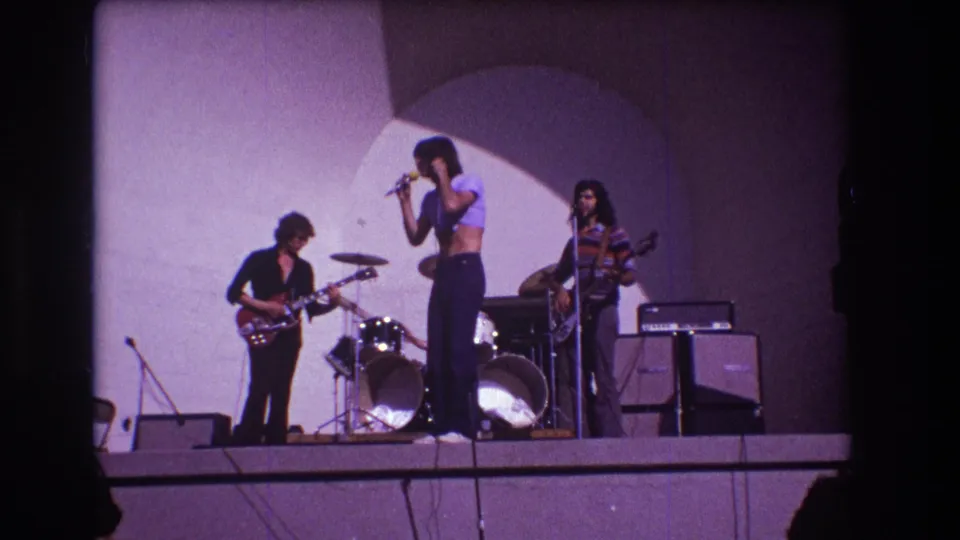 The 1973 Medford stage view holds on the singer at center, with guitarist, bassist, drum kit, microphone stands, and amplifier cabinets visible.