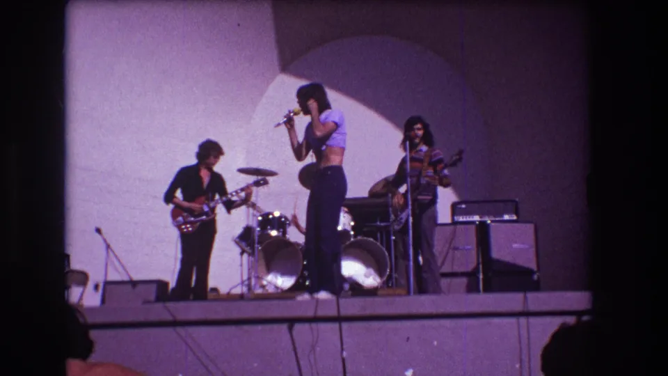 In this Medford 1973 frame, the vocalist shifts posture while the guitarist and bassist remain playing beside the drum kit and stage equipment.