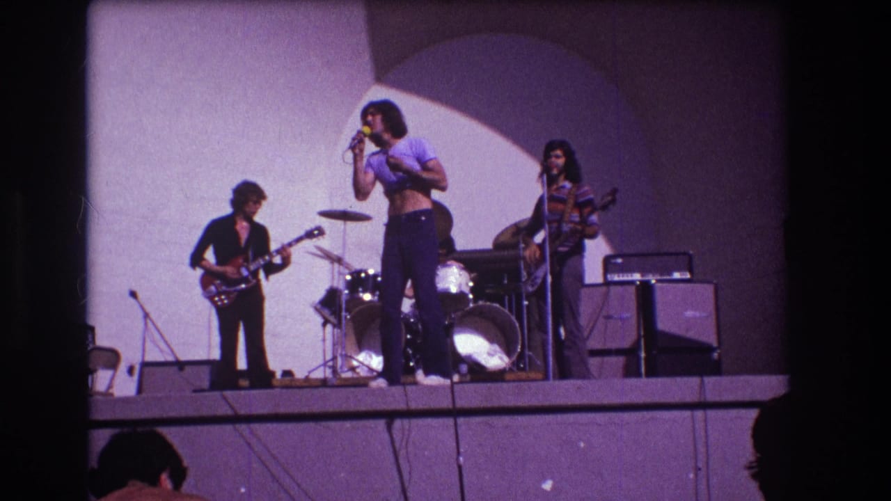 1973 Medford, New Jersey 8mm frame showing a rock band onstage with vocalist at microphone, guitarist, bassist, drums, and amplifiers.