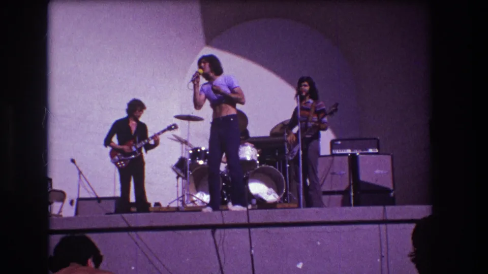 1973 Medford, New Jersey 8mm frame showing a rock band onstage with vocalist at microphone, guitarist, bassist, drums, and amplifiers.