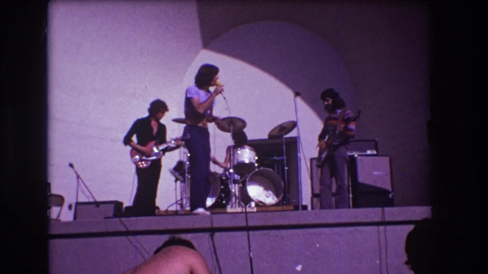 1973 Medford, New Jersey 8mm frame with singer facing the bassist, guitarist at left, drum kit behind, and amplifiers at right.