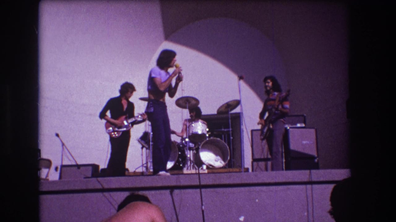 1973 Medford, New Jersey home-movie frame of a four-piece rock band performing on an outdoor stage under an arched backdrop.